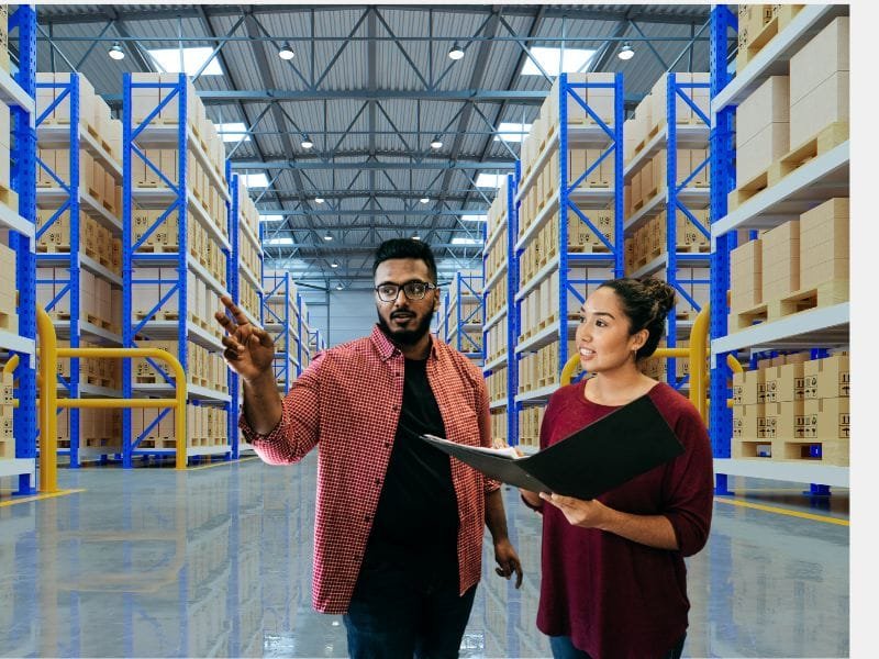 A man in a red shirt and a woman in a burgundy top discuss inventory in a large warehouse filled with stacked boxes.